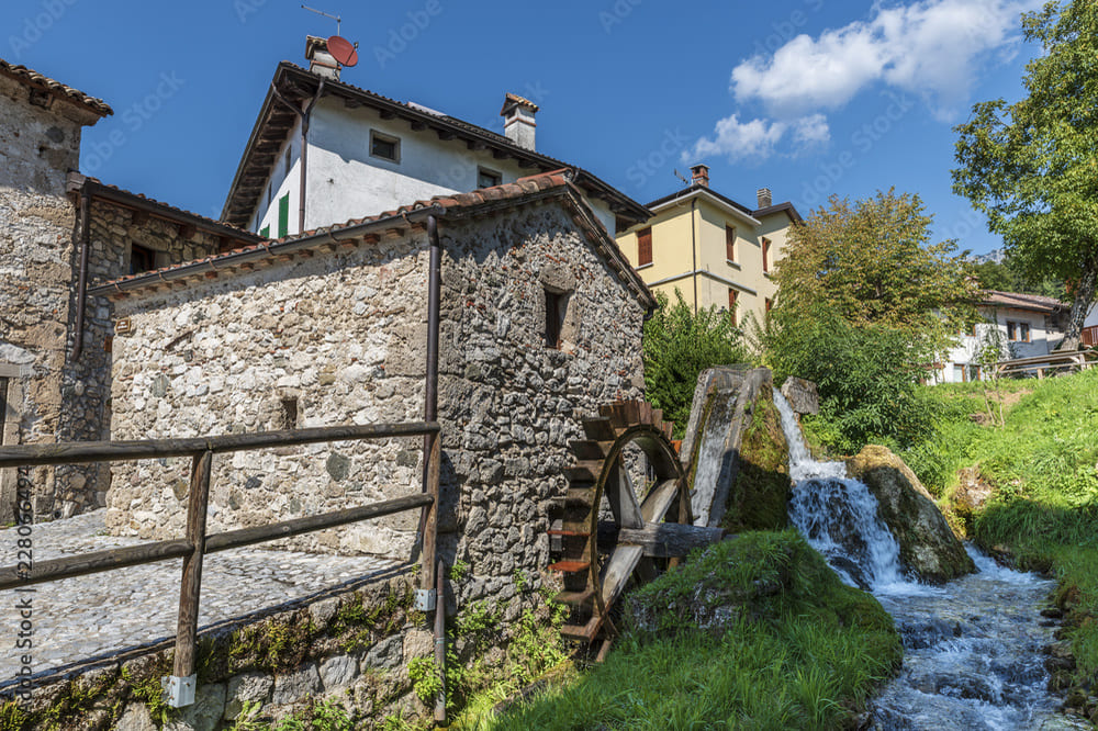 Mulino ad acqua storico lungo la Via dei Mulini a Illegio, caratterizzato da una piccola cascata e una ruota in legno, testimone della tradizione agricola del borgo.