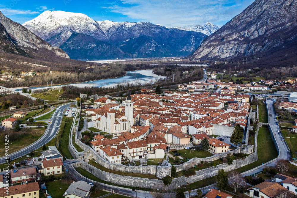 Vista aerea panoramica di Venzone, mostrando il centro storico circondato da mura medievali, con le verdi colline e i corsi d'acqua in lontananza.