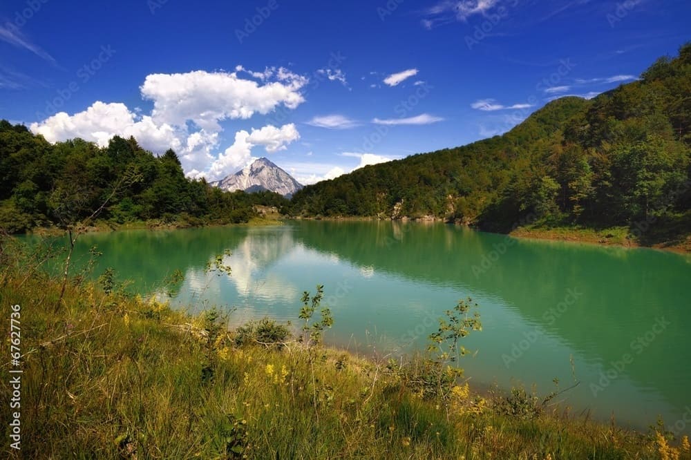 Vista panoramica del Lago di Verzegnis, circondato da vegetazione lussureggiante e con il Monte Amariana in lontananza.